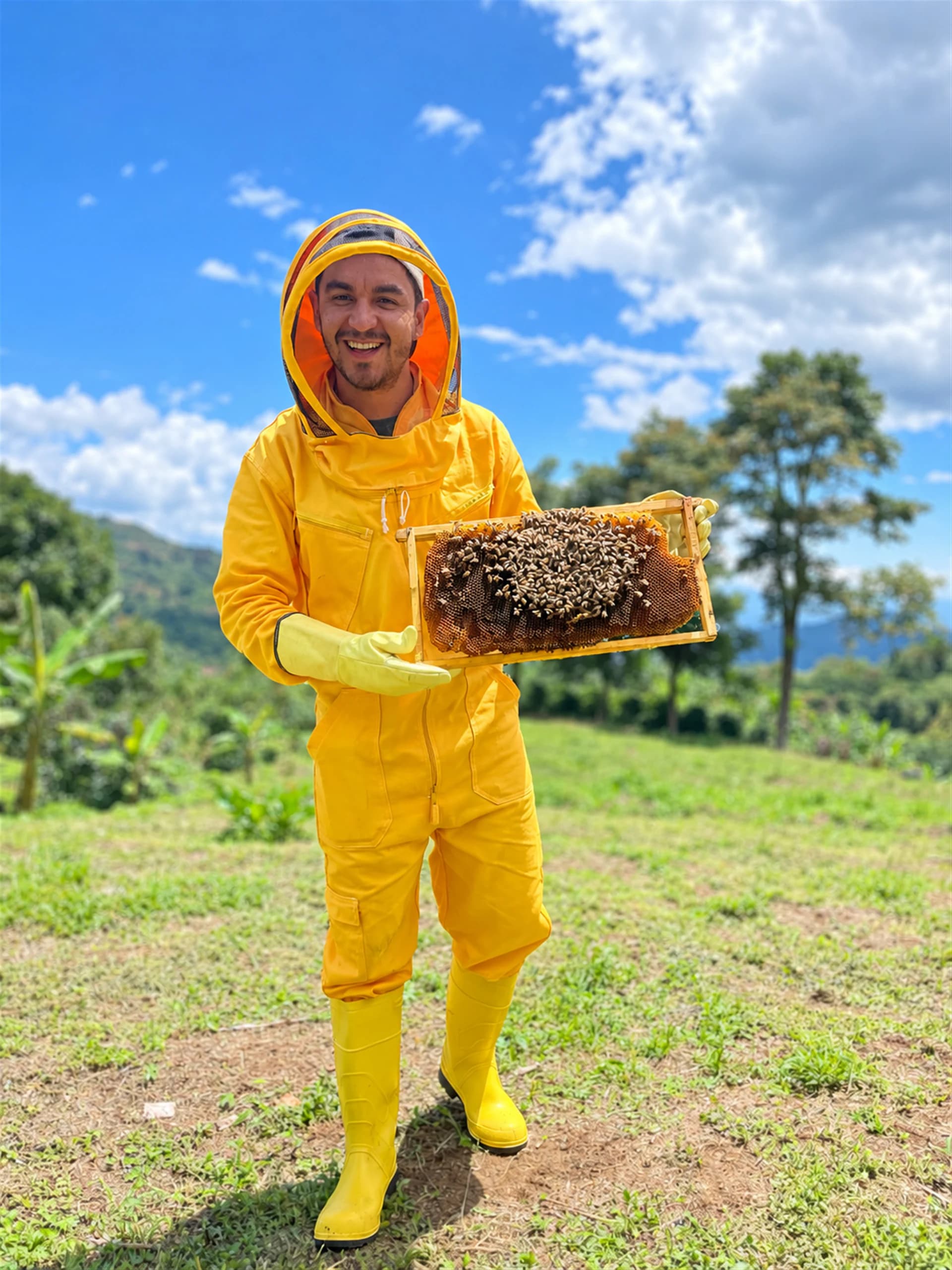 A beekeeper in a yellow suit holds a honeycomb frame in a lush green landscape.