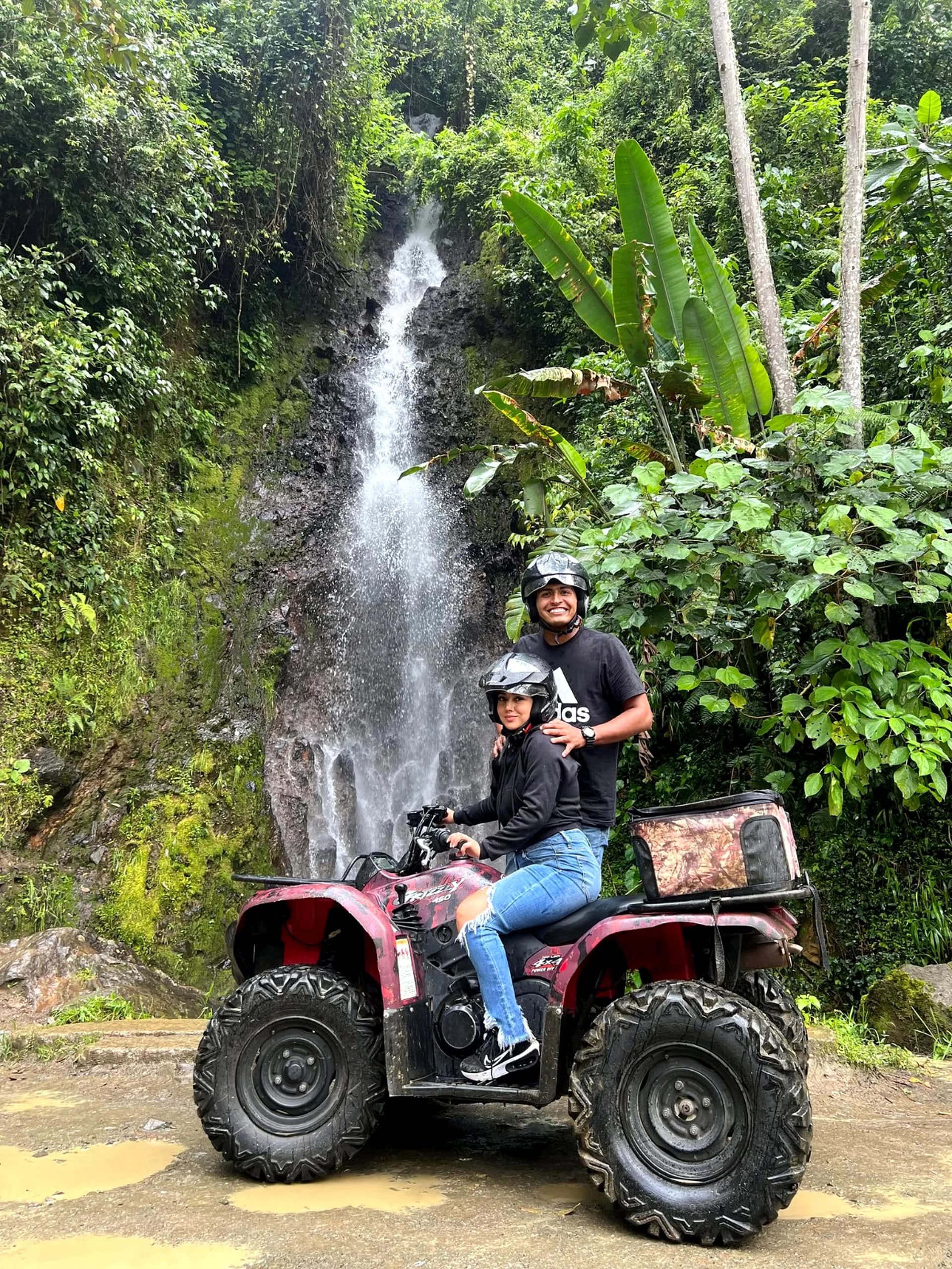 Pareja sonriente sobre cuatrimoto frente a una cascada en Jardín, Antioquia.