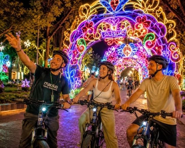 Three people on electric bikes under an arch of Christmas lights in Medellín.